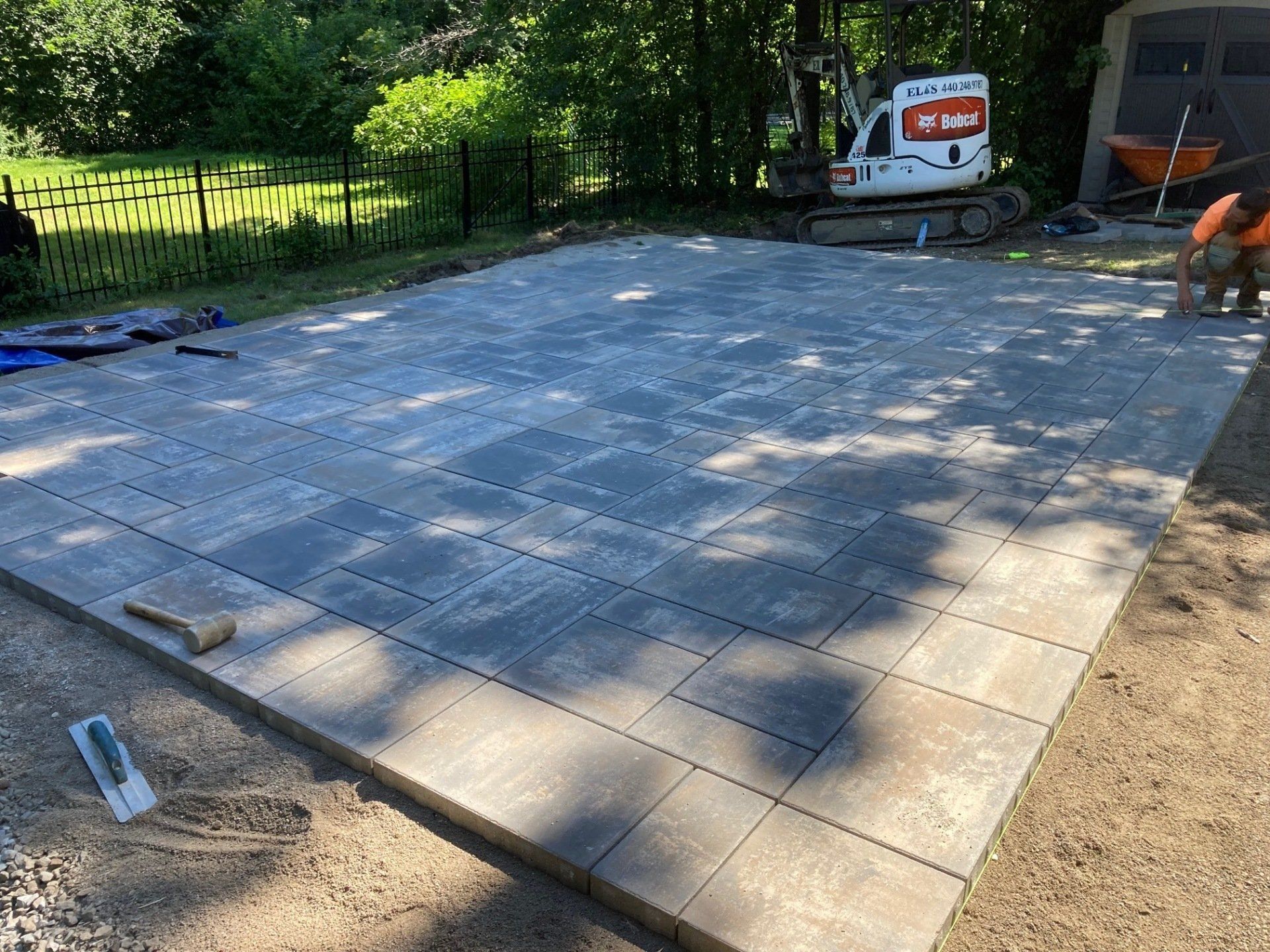 Outdoor patio under construction with gray pavers, tools, and a small excavator nearby.