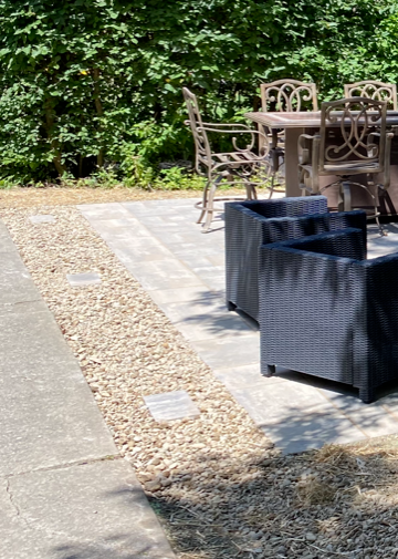 Outdoor patio with wicker chairs, table, and planters beside a gravel strip and concrete walkway.