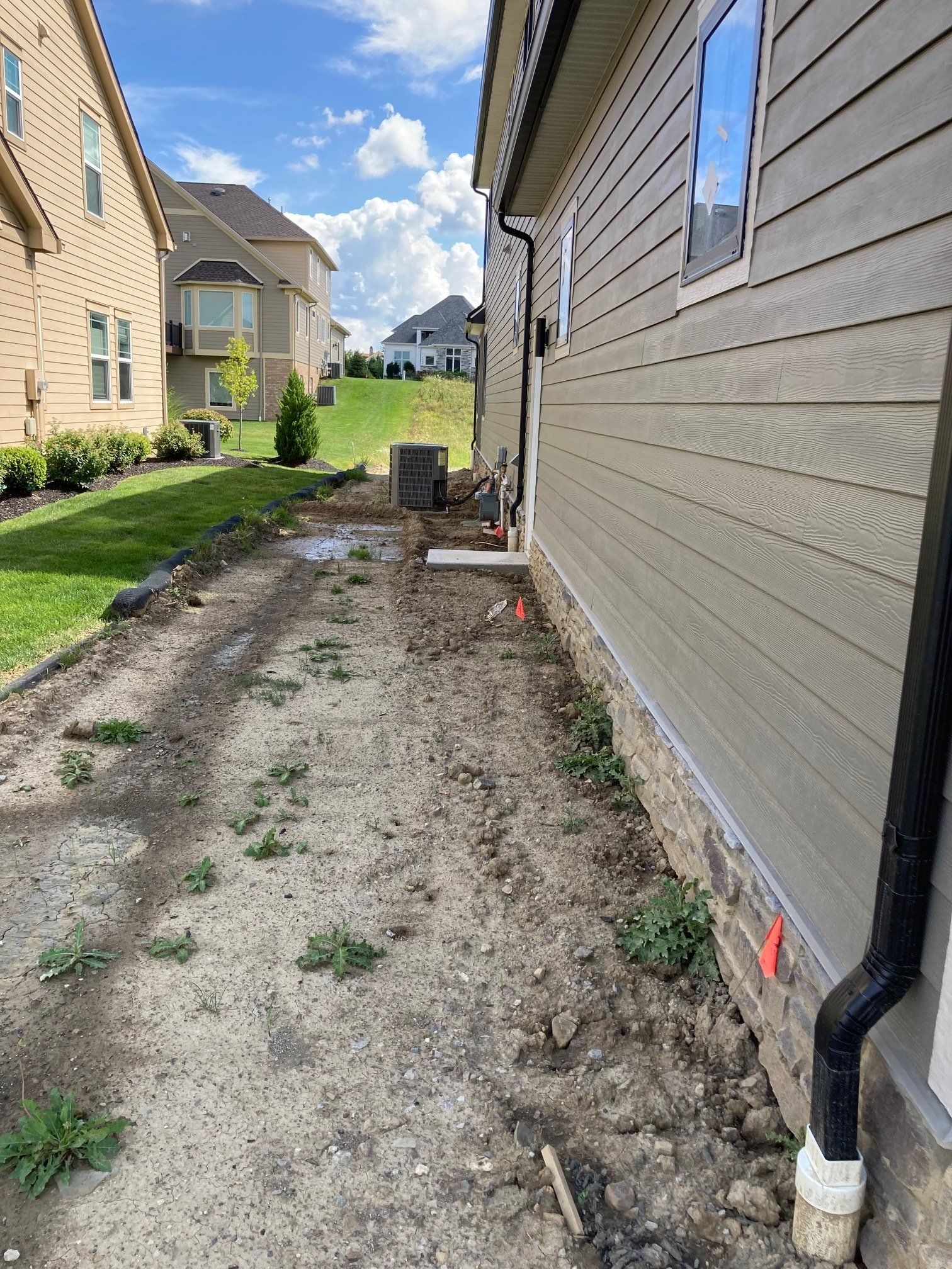 Narrow gravel side yard between houses with a black drainpipe and a white vent along the right wall