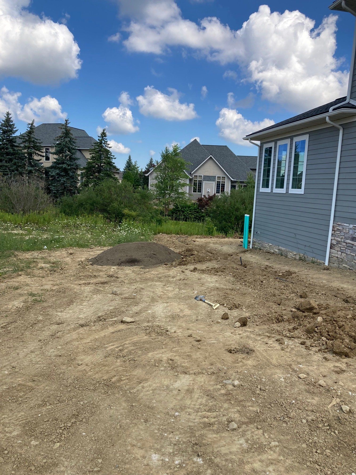 Dirt yard beside a gray house under a blue sky, with trees and mountains in the background