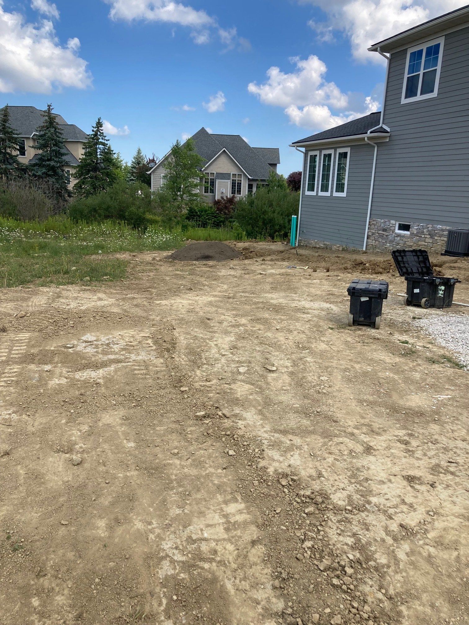 Dirt lot beside a gray house under blue sky, with trash bins and neighboring homes in the background