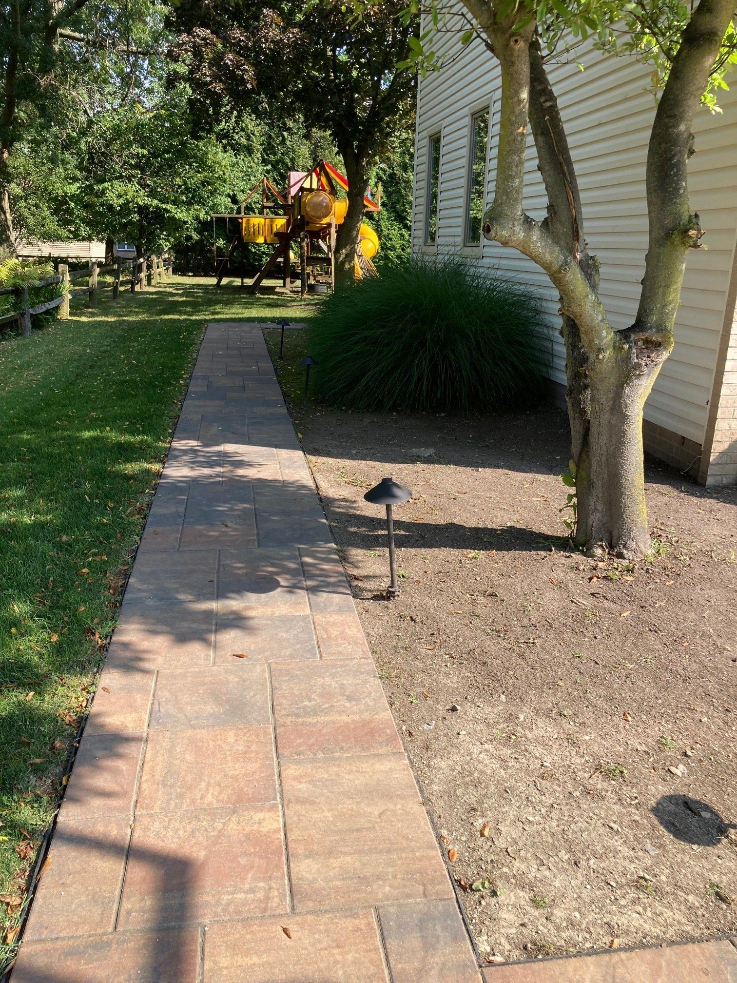 Garden walkway beside a house, with trees, shrubs, and a person in a yellow shirt in the distance