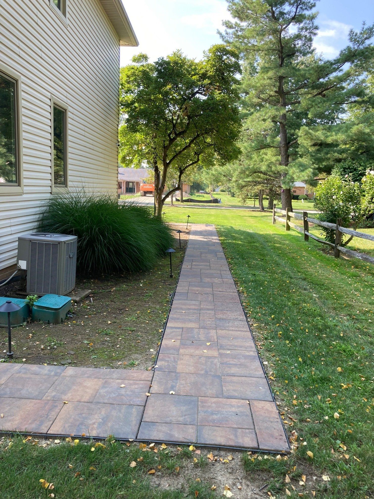 Brick walkway beside a house leading through a grassy yard with trees and a bench