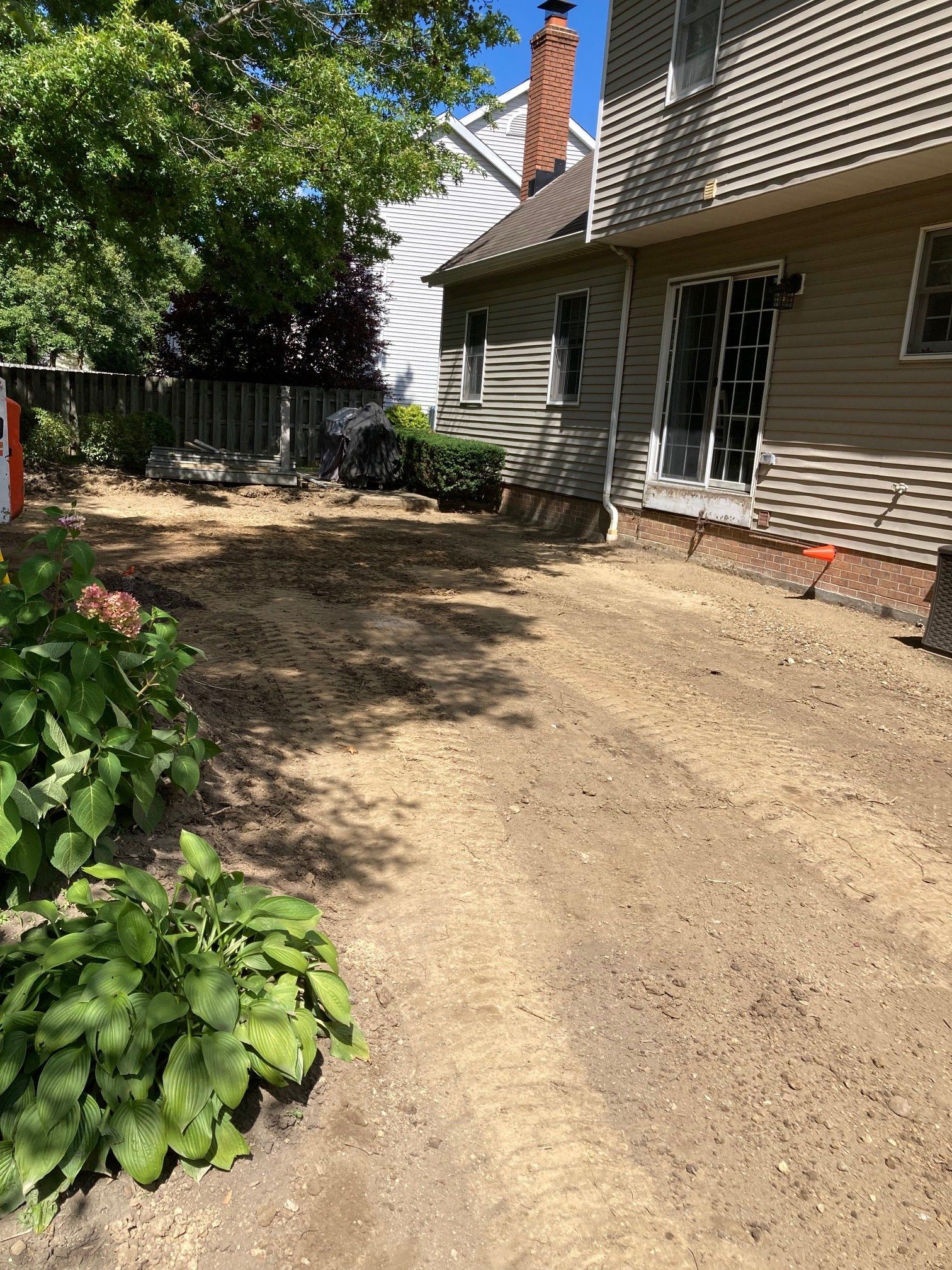 Backyard beside a house, with a dirt path, green plants, and a brick chimney under leafy trees.
