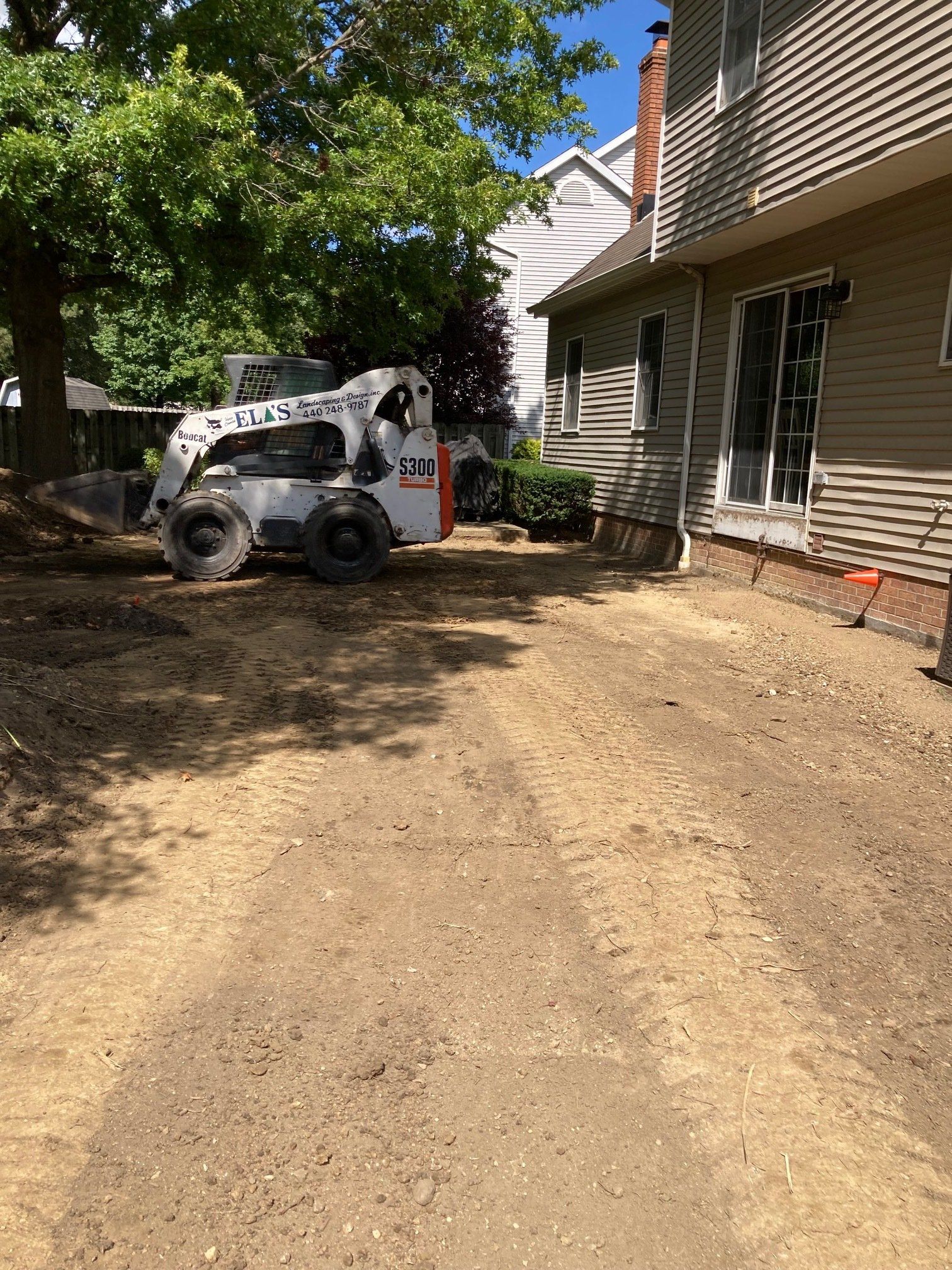 White skid steer on dirt driveway beside house under trees, with exposed siding on the right.