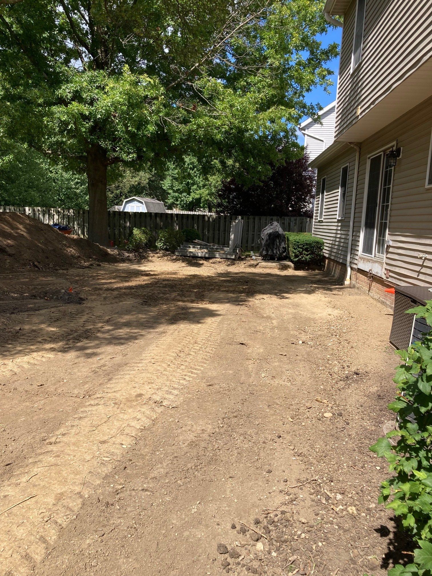Backyard dirt path beside a house, with trees and a fence in the background.
