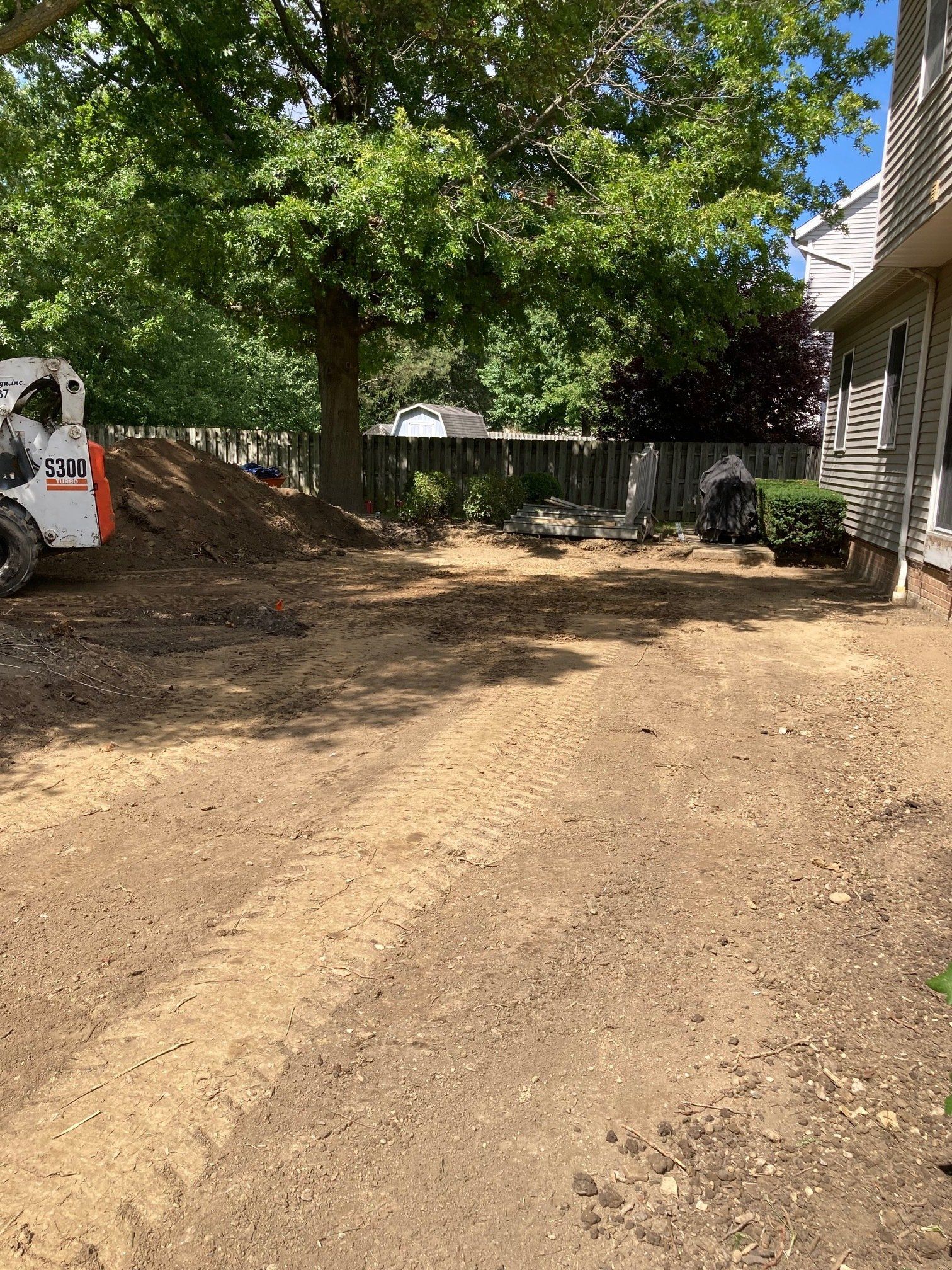 Dirt driveway beside a house and trees, with a white vehicle at left and a fence in the background.