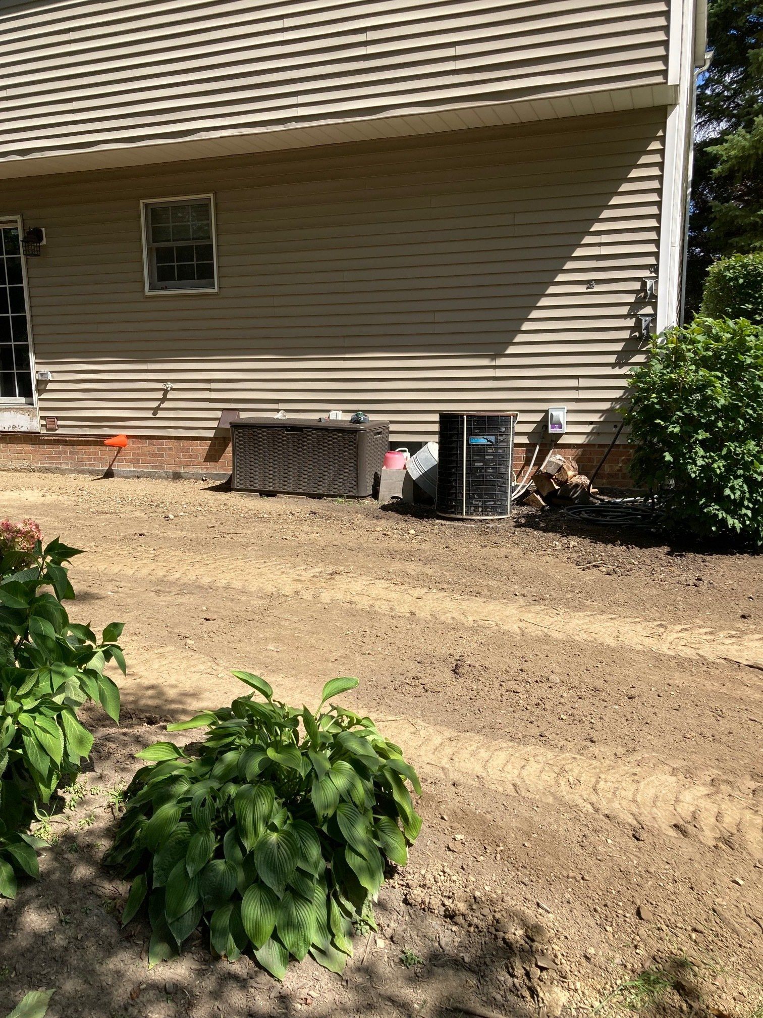 House side yard with fresh dirt, shrubs, and outdoor equipment against beige siding