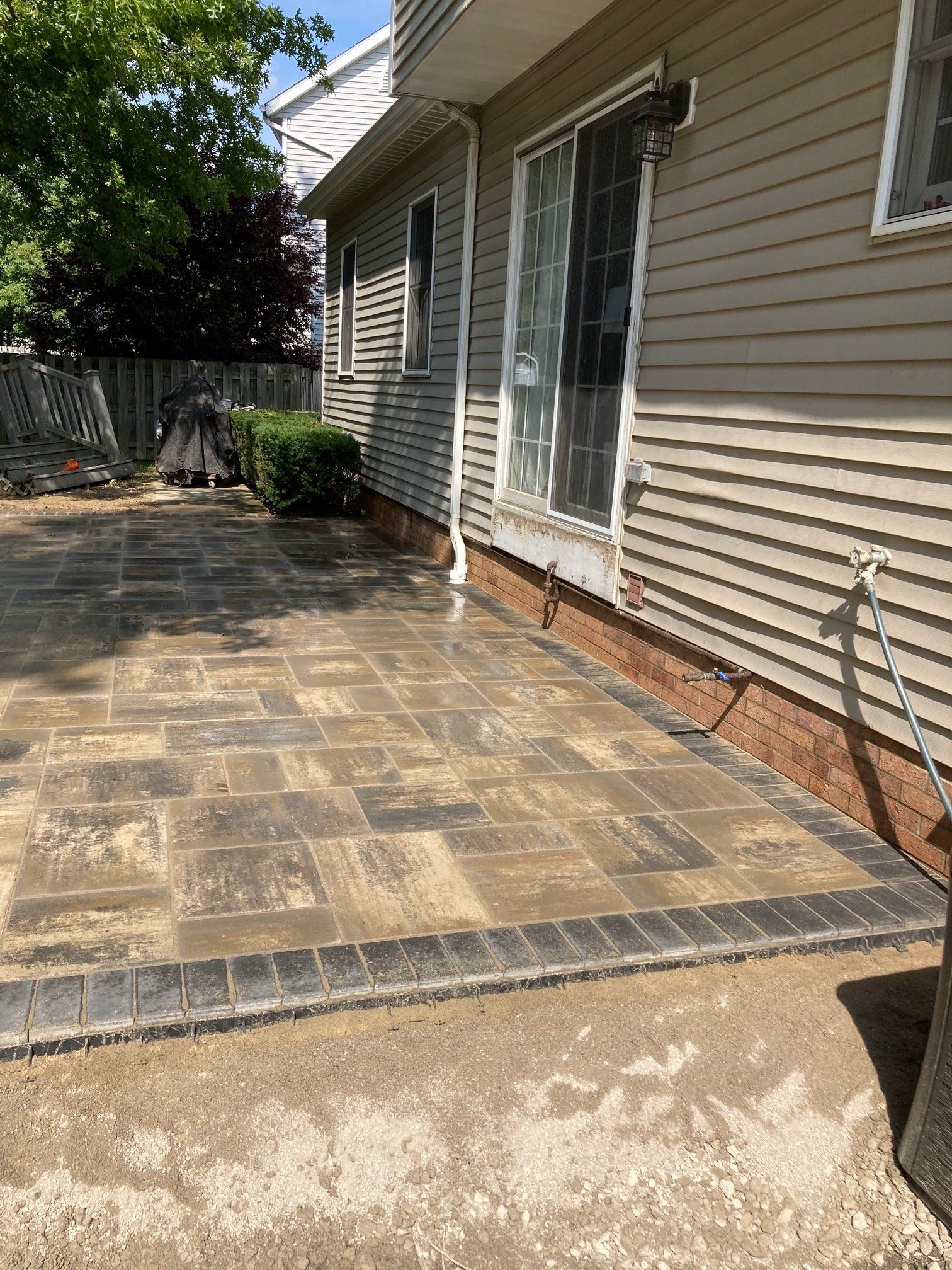 Stone patio beside beige house with sliding glass doors, bordered by shrubs and a wooden fence.