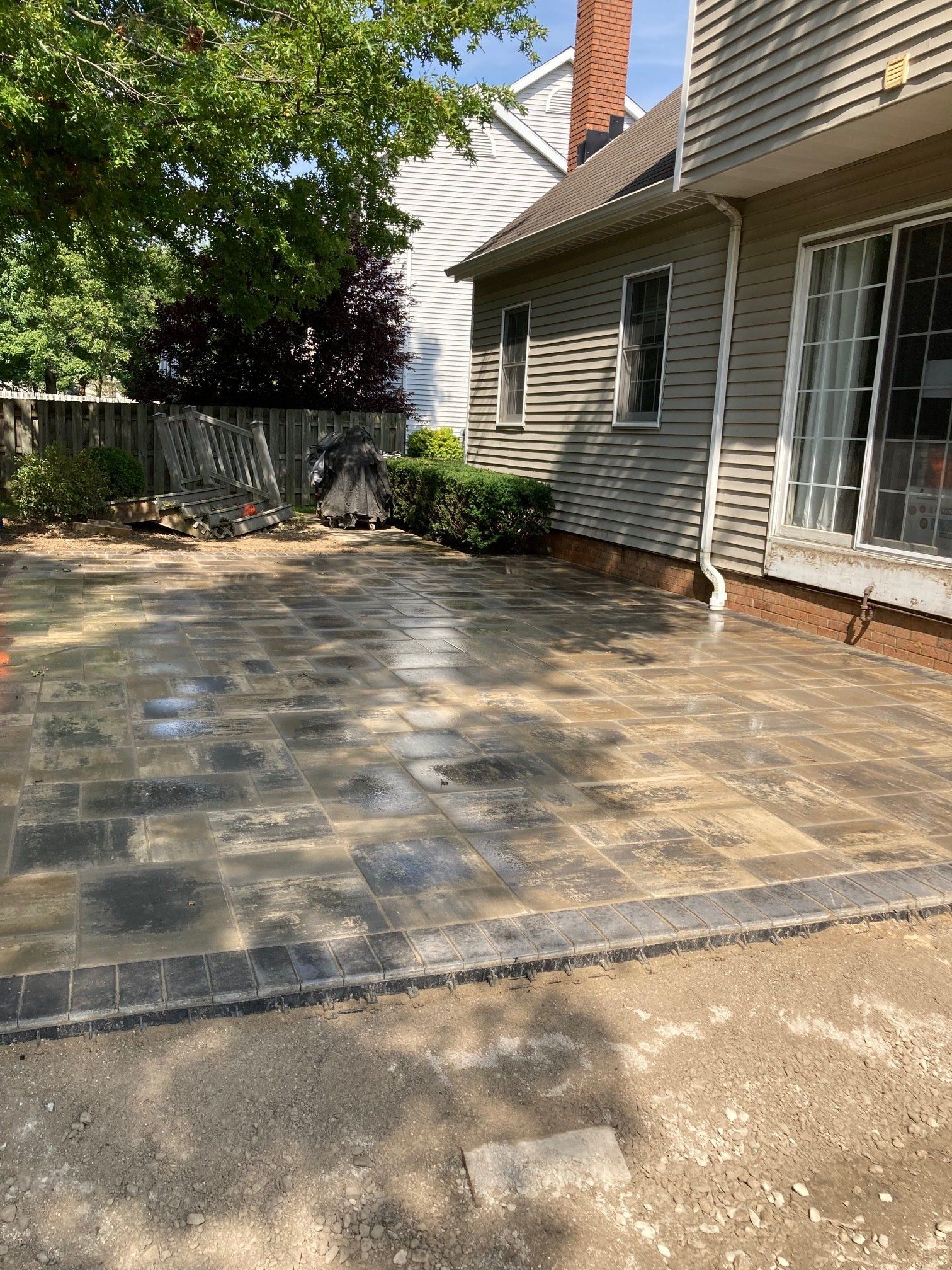 Patio with wet stone pavers beside a house, bordered by a fence and trees in sunlight