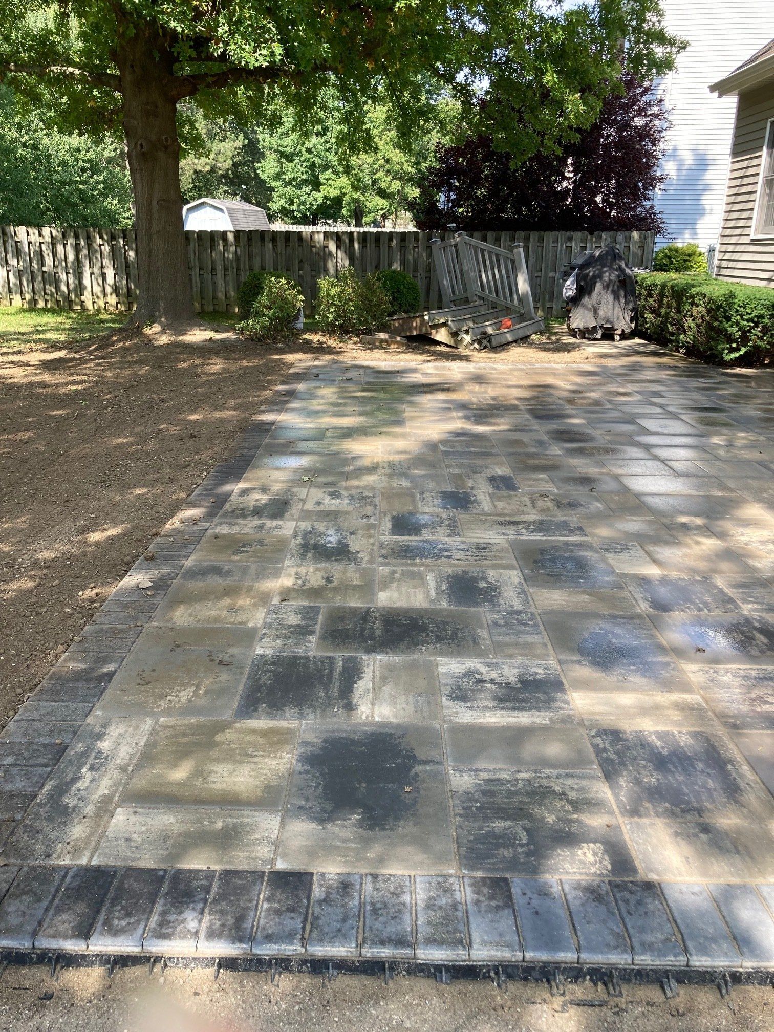 Paved stone patio in a backyard with trees, fence, and house siding in the background