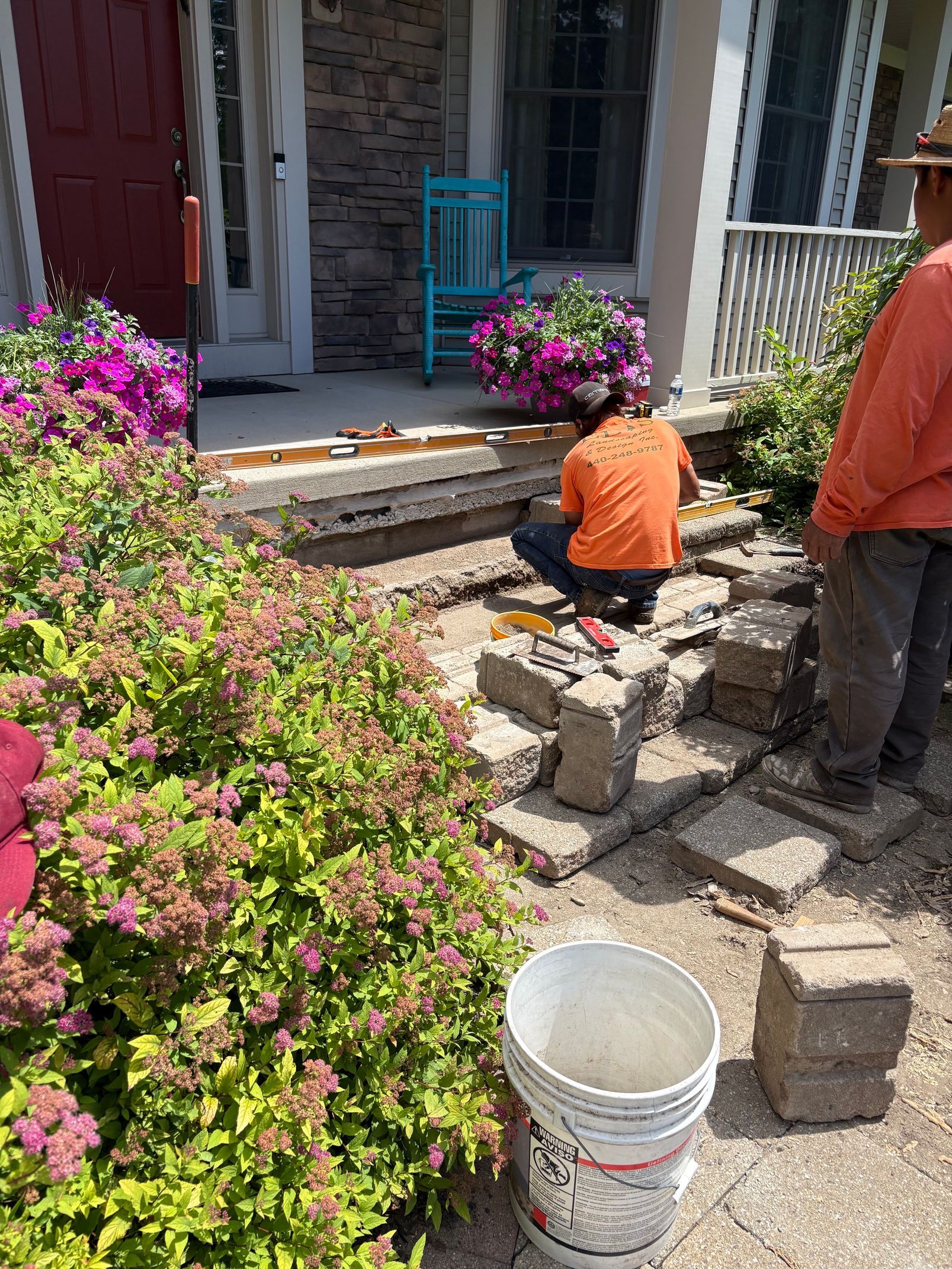 Two workers building a stone walkway beside a house, with pink flowers and a bucket in the foreground.