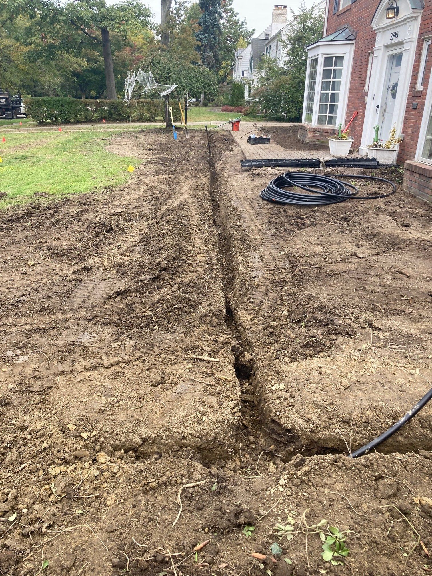 Freshly dug trench across a bare yard beside a red-brick house, with a hose and coiled cable nearby.