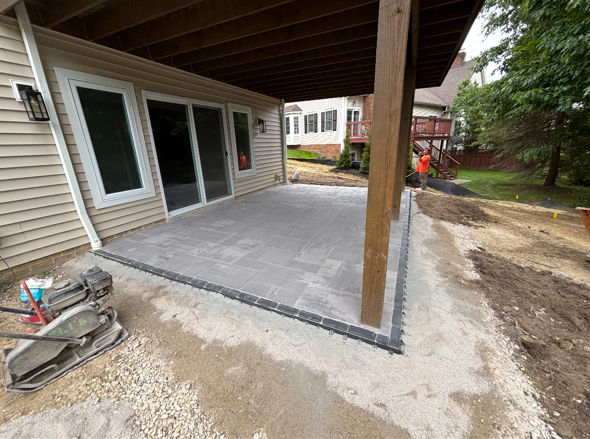 Covered concrete patio under construction beside a house, with a wooden support post and unfinished edges.