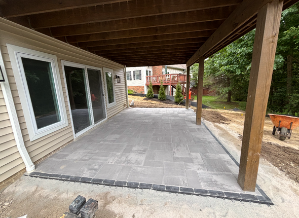 Covered patio with gray pavers, sliding glass doors, and wooden posts beside a house