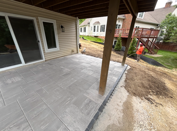 Concrete patio under covered porch beside house, with a worker in orange jacket near the yard.