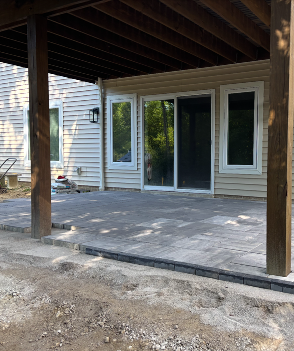 Covered concrete patio with sliding glass doors and dark wooden support posts under a roof.