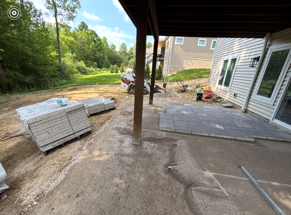 Backyard patio under construction with stacked pavers, dirt ground, and a house exterior.