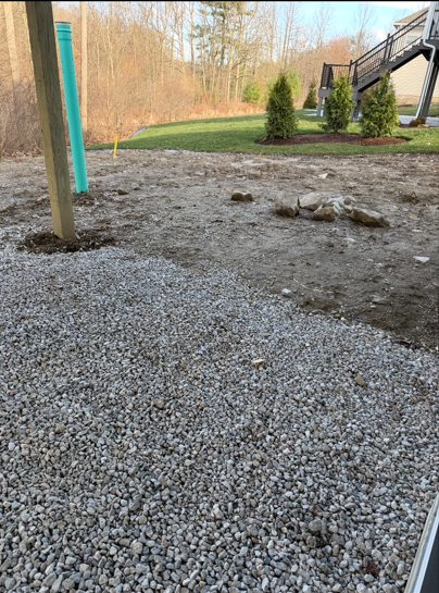 Gravel yard with a patio post, dirt patch, shrubs, and trees in the background