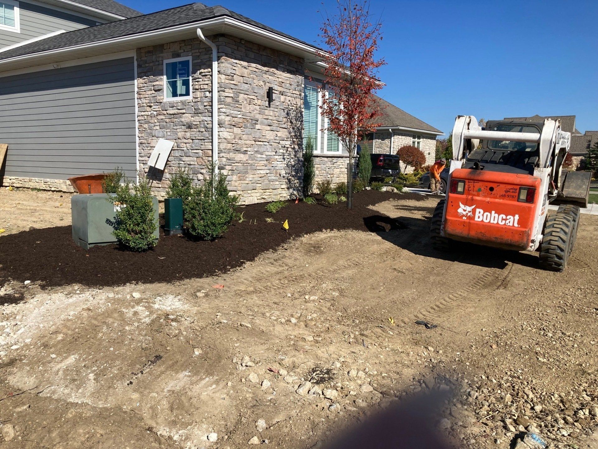 Construction site beside a house, with a Bobcat loader on a gravel driveway and landscaping in progress