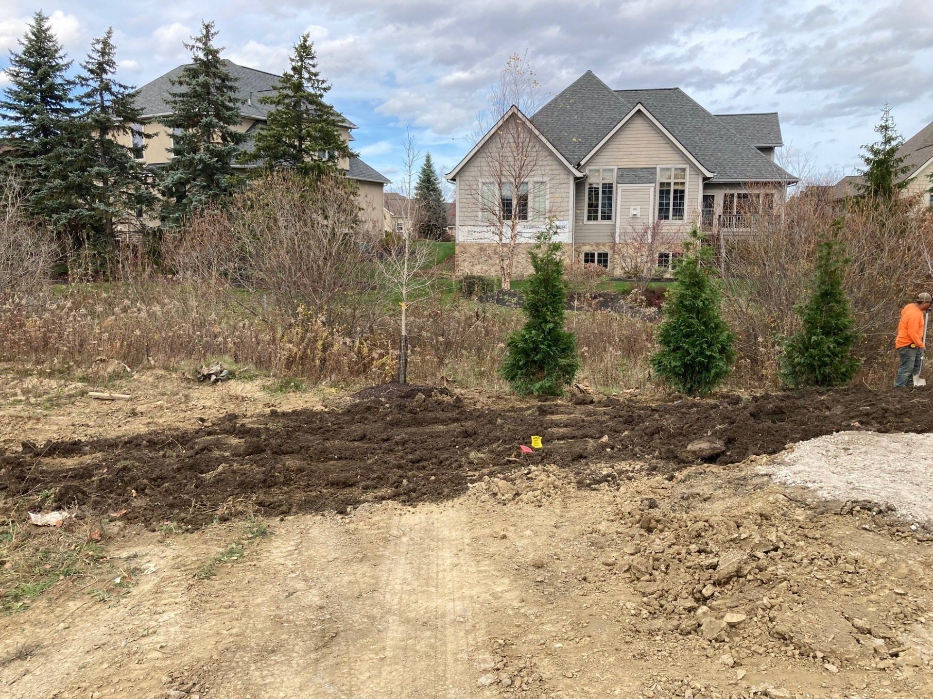 Dirt lot with a beige house and evergreens; a person in orange stands at right.
