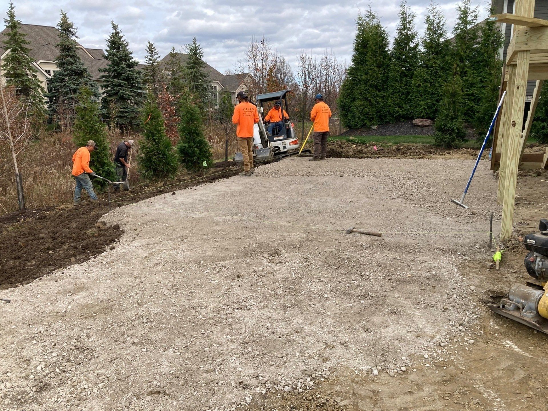 Workers in orange vests landscaping a gravel yard beside evergreen trees