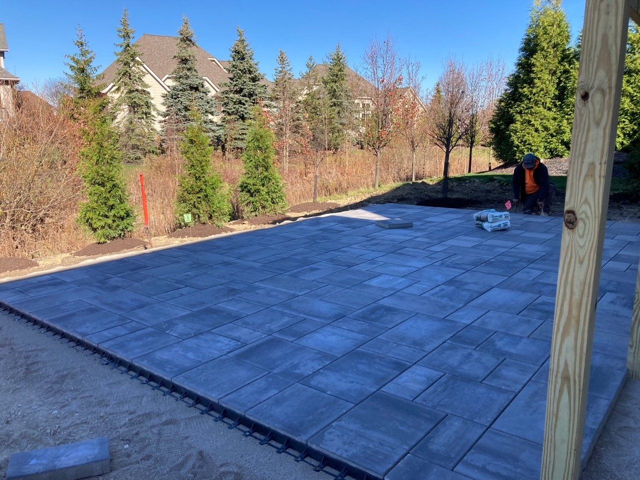 Backyard patio with gray pavers, trees and shrubs along the edge, and a worker near the far side.