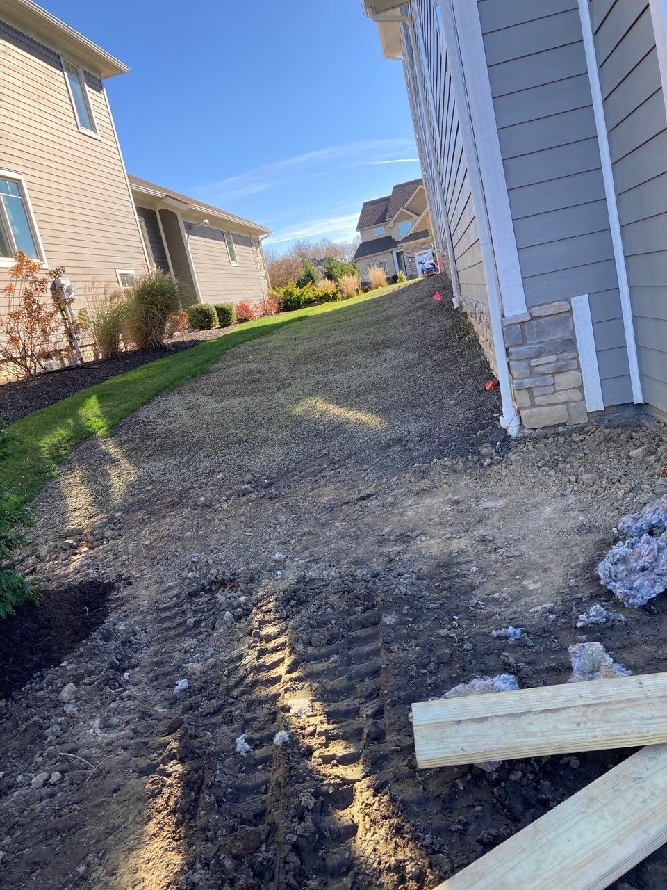 Narrow gravel path between two houses under a clear blue sky, with construction debris in the foreground