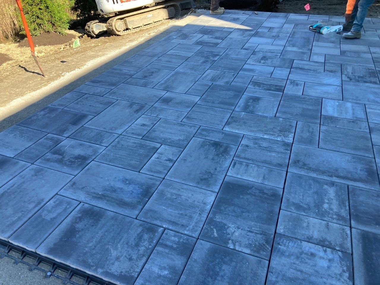 Freshly laid gray stone pavers forming a driveway or patio edge near a curb and workers' legs