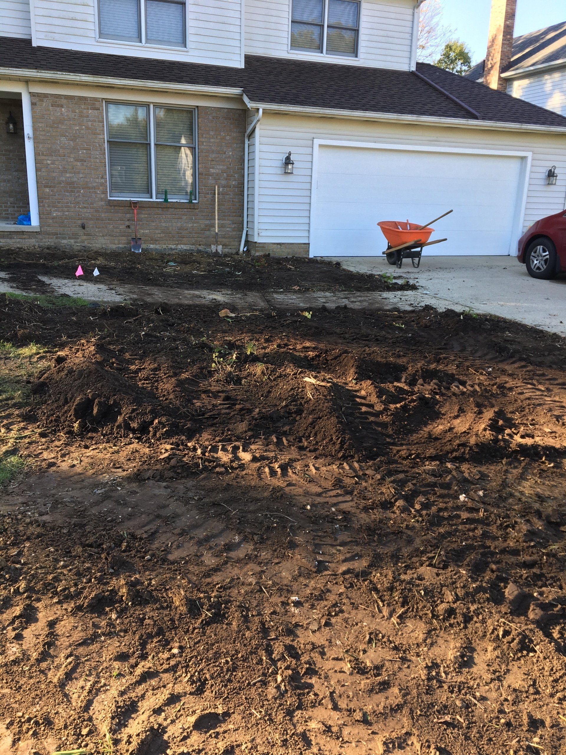 Front yard under renovation with bare soil, a wheelbarrow, and a brick house with a white garage.