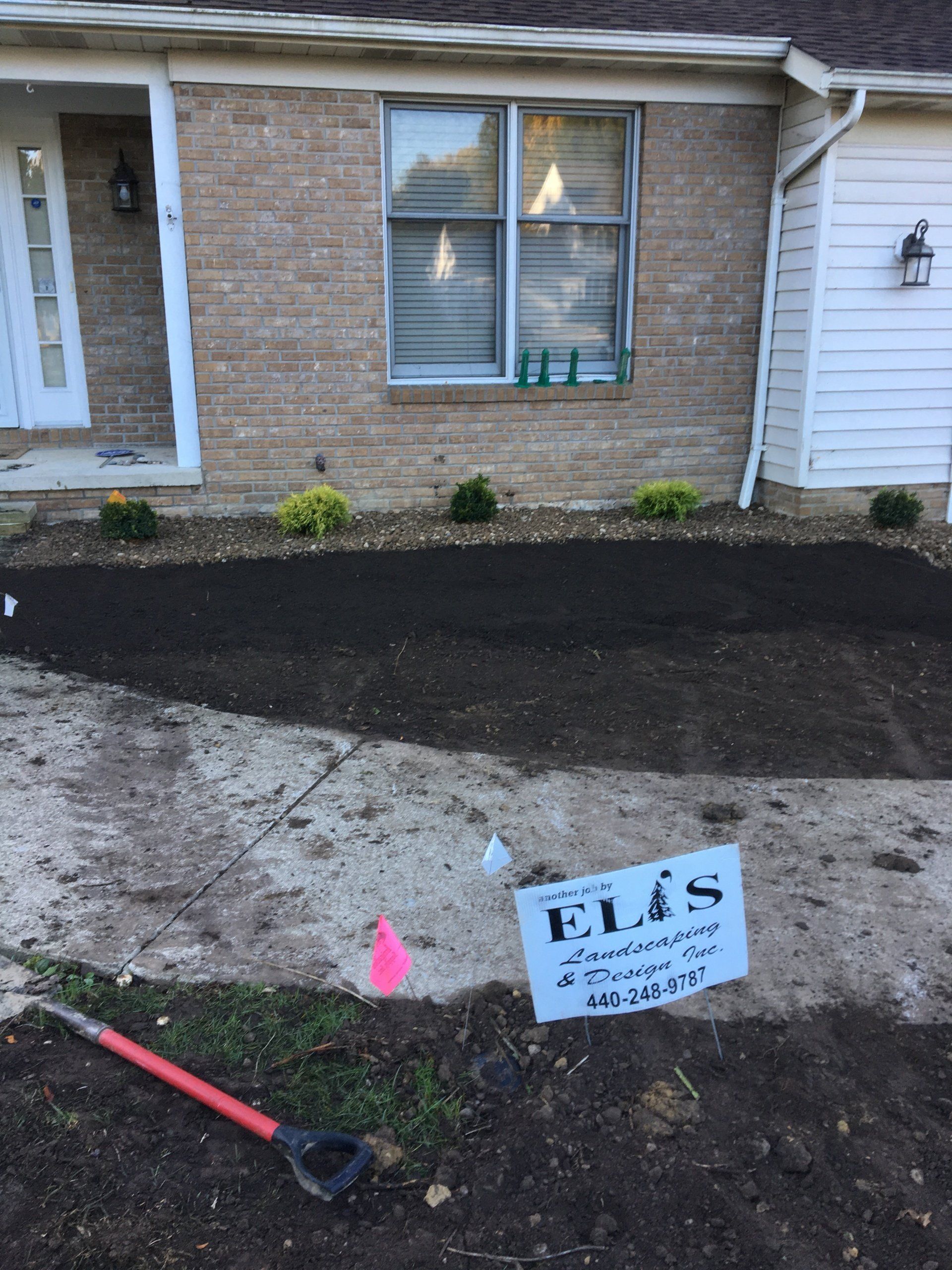 Front yard mulch bed by brick house, with black soil, a shovel, and a small E.L.S. sign.