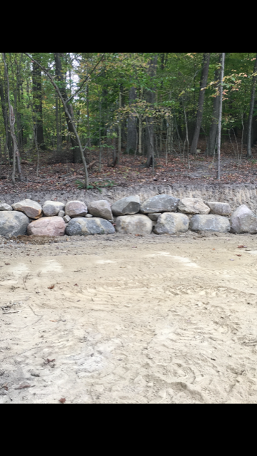 A stone retaining wall borders a sandy path with a wooded area in the background.
