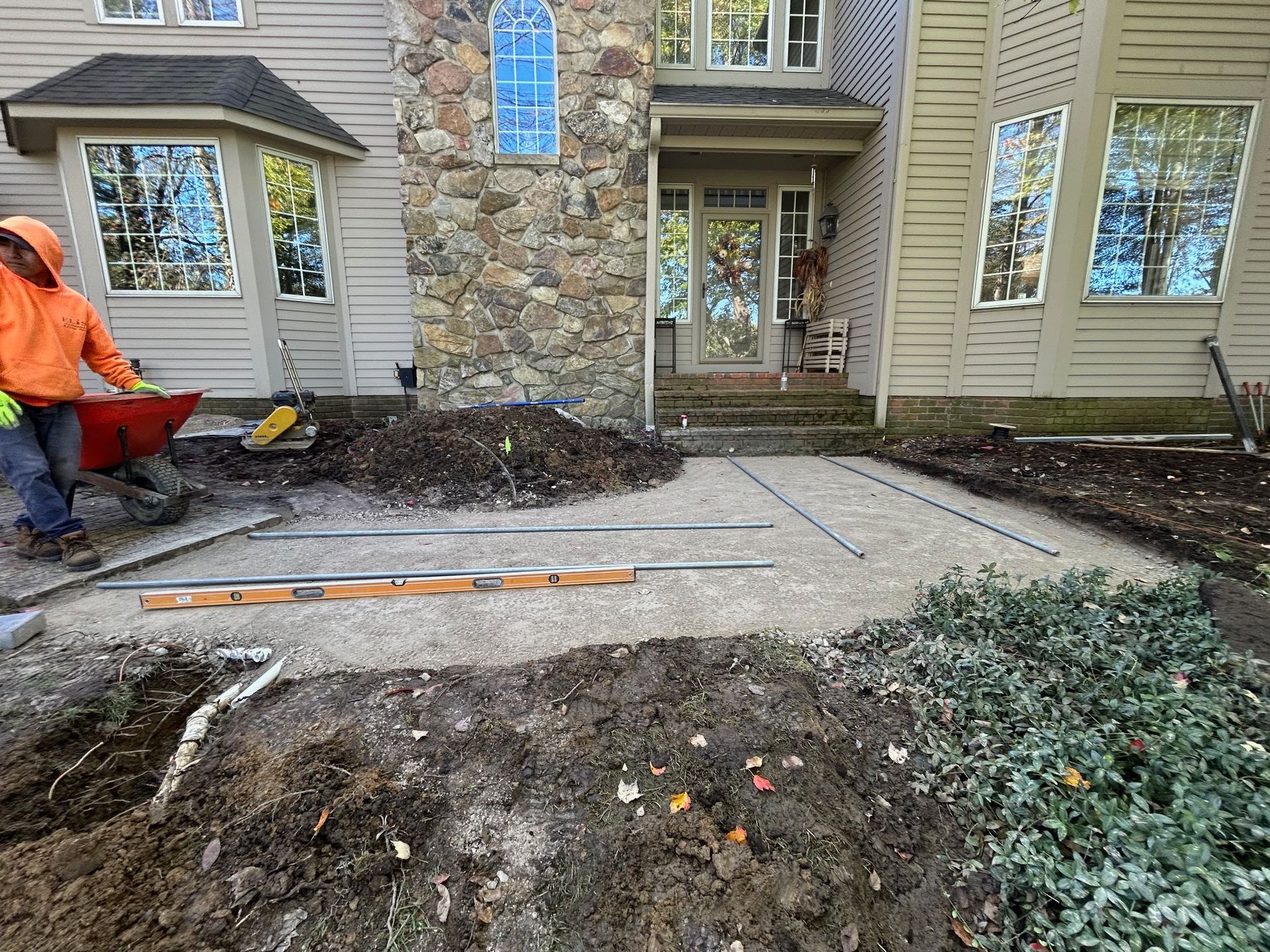 Worker in orange jacket installing a concrete walkway beside a stone-front house and garden beds