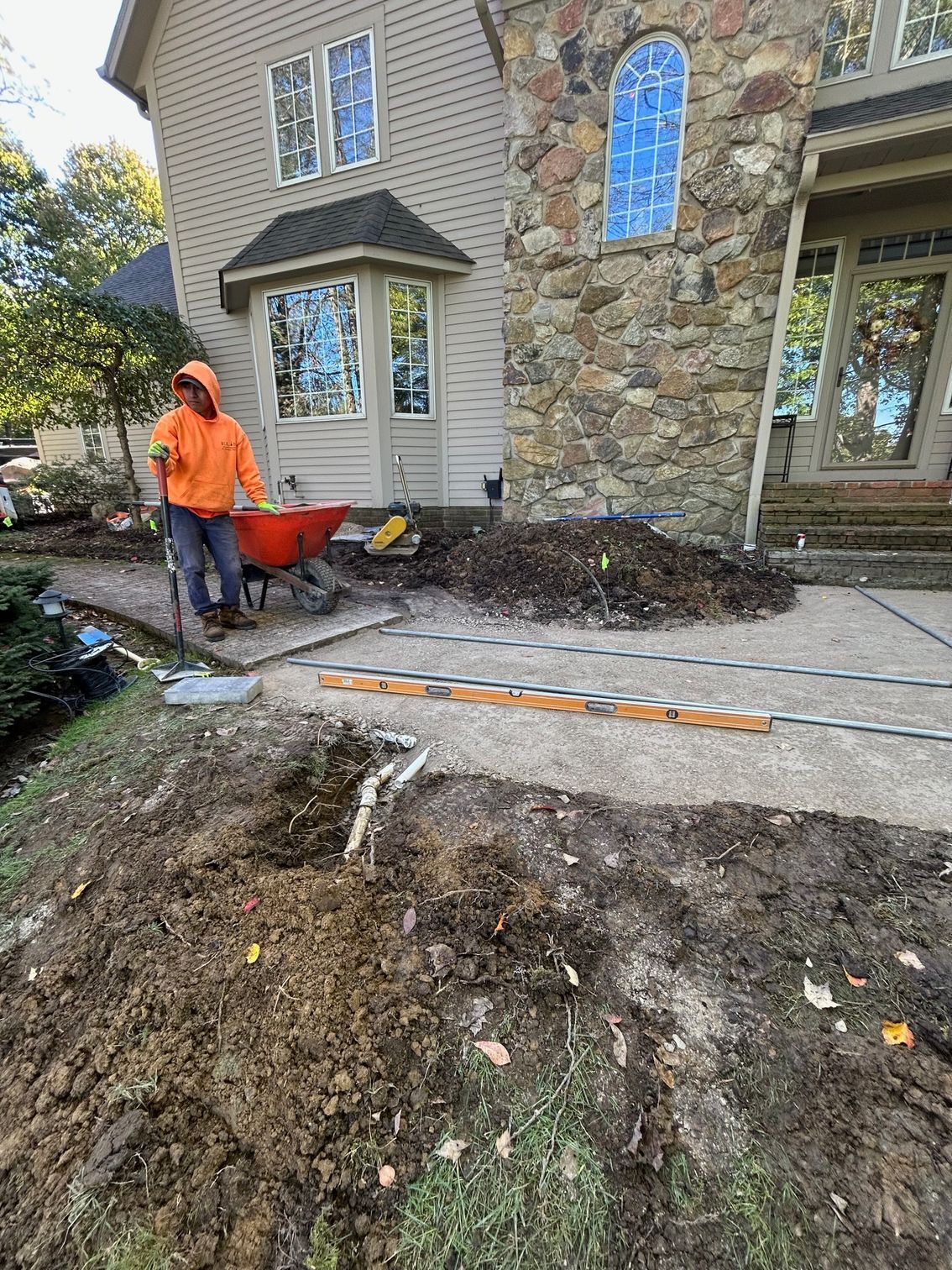 Worker in orange jacket with wheelbarrow beside a stone house and muddy front yard during landscaping.