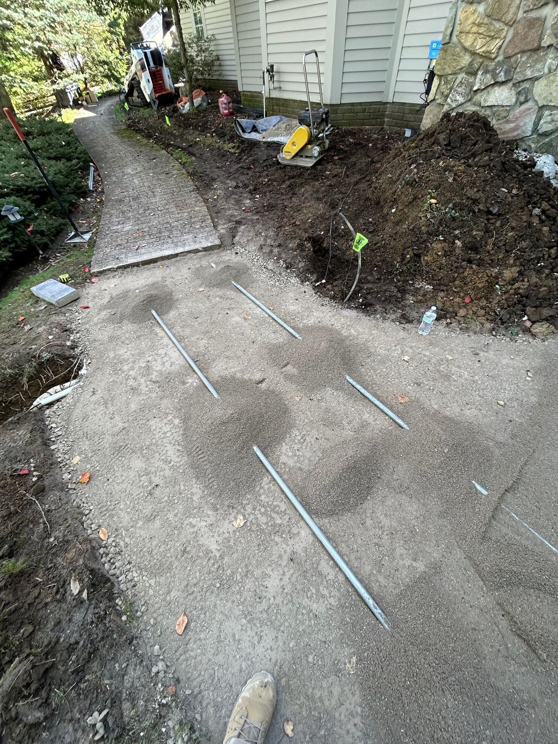 Sidewalk under repair beside a house, with dug-up soil, tools, and workers in the background.