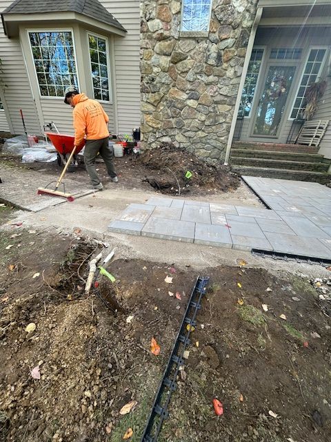 Worker in orange shirt raking soil beside a house walkway and stone wall