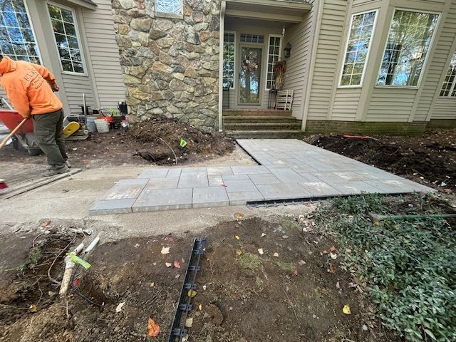 Worker paving a concrete walkway beside a stone house and landscaped yard