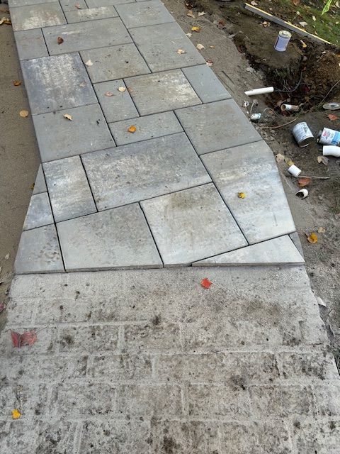 Sidewalk with light gray paving stones beside a dirt edge and scattered litter near a curb cut.