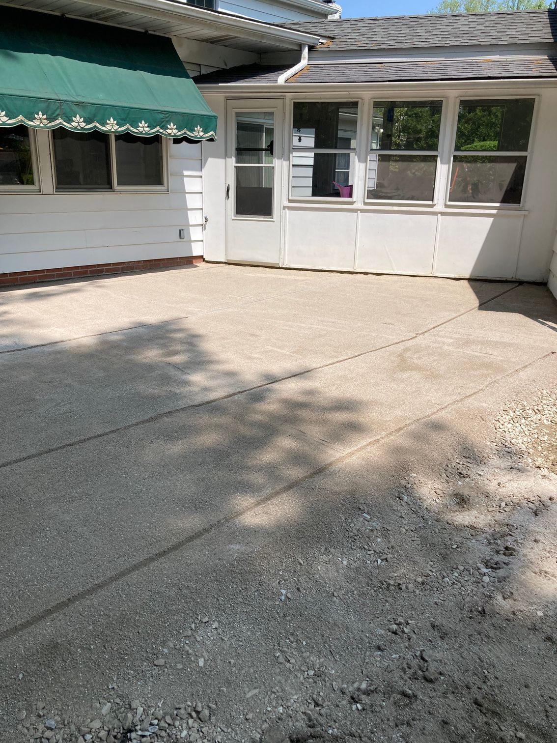 Empty concrete courtyard beside a white house with green awnings and a sunlit screened porch.