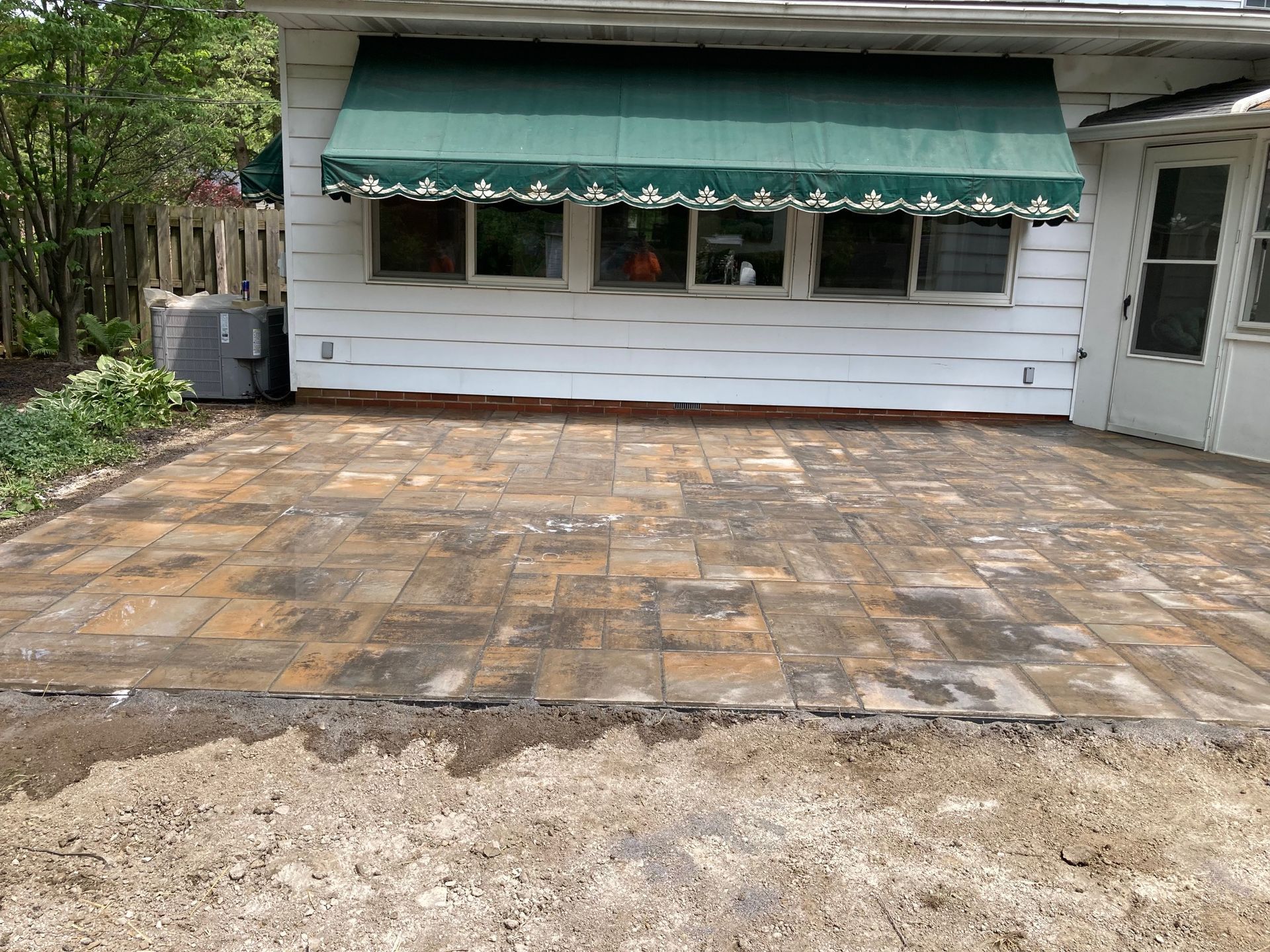 Stone patio in front of a white house with a green awning and small windows, beside a gravel yard.