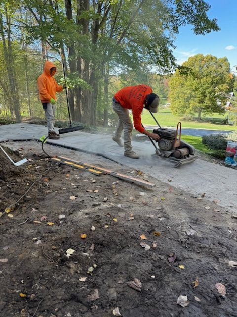 Workers in orange jackets paving a path with a plate compactor beside a wooded park area