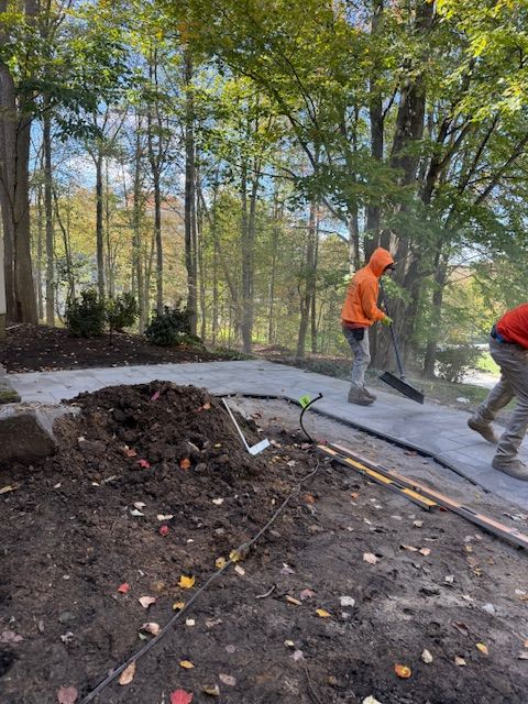 Workers in orange hoodies move dirt near a paved path in a wooded area.