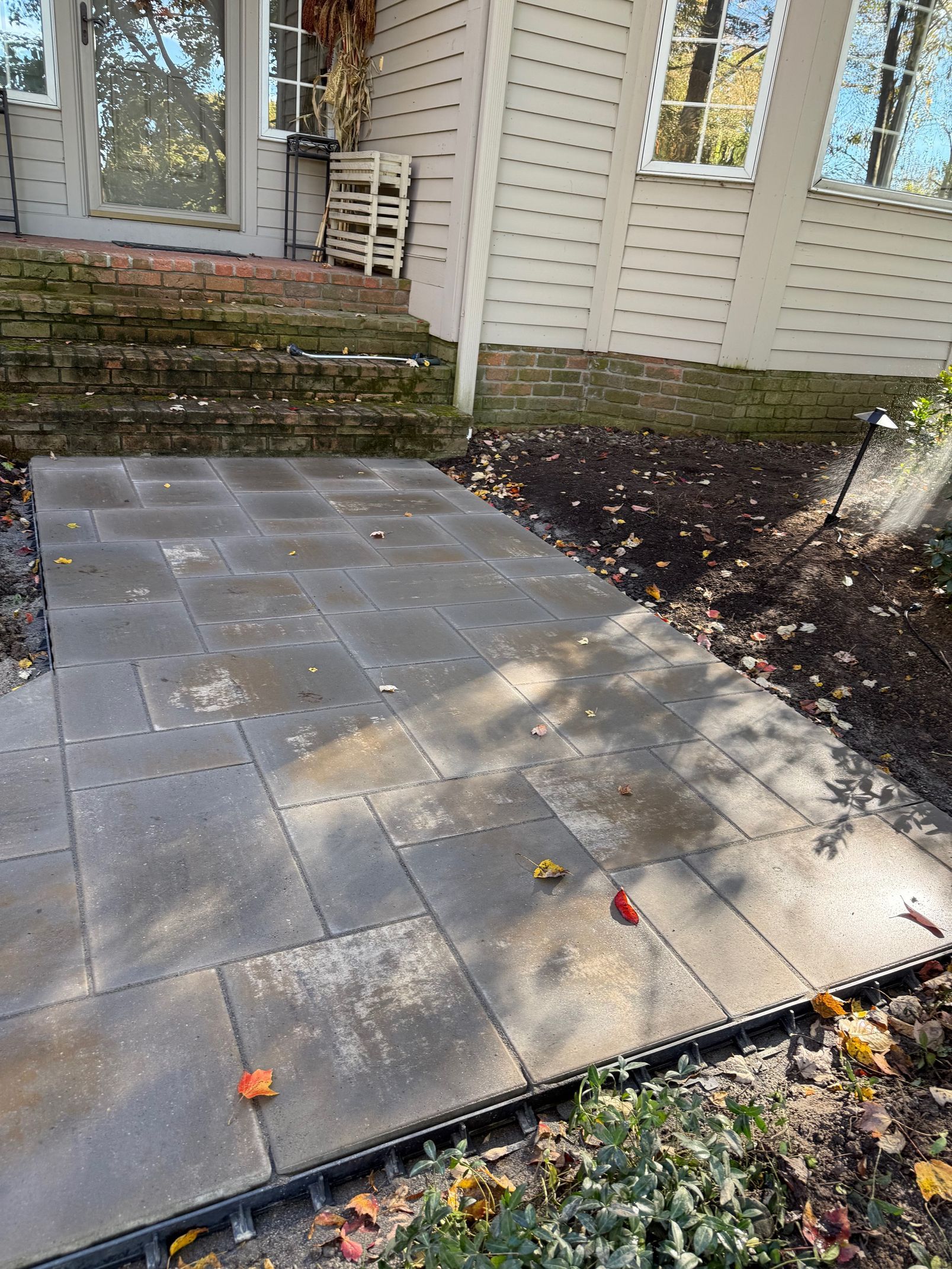 Patio with gray pavers beside a house, bordered by plants and fallen leaves in sunlight