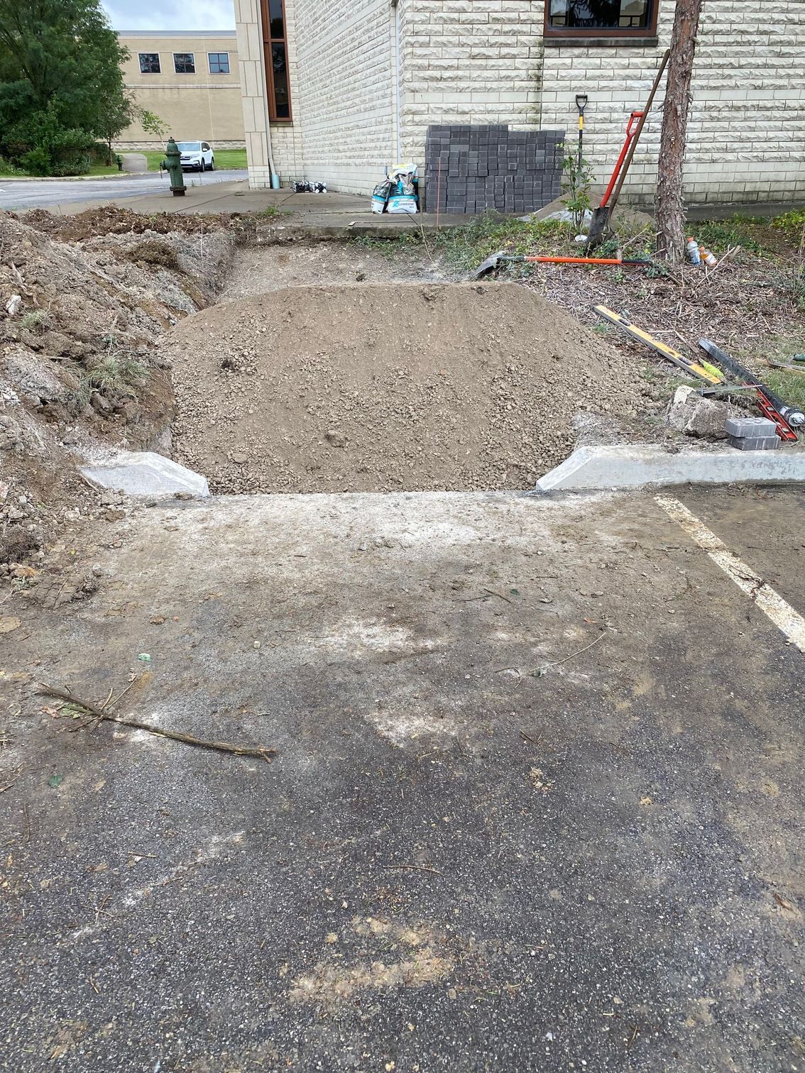 Large dirt excavation in a parking lot beside a brick building, with concrete edges and a shovel nearby