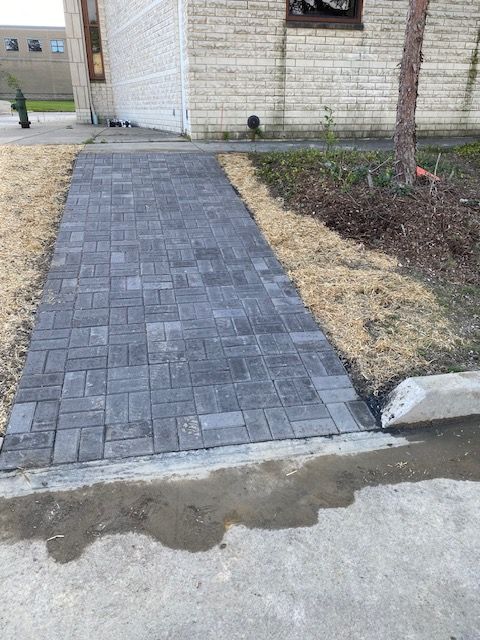 Brick paver walkway leading to a house entrance beside a concrete drive and landscaped bed