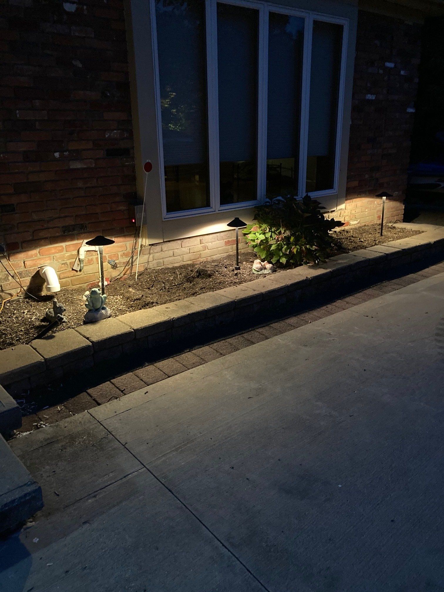 Dimly lit front walkway beside a brick house, with landscape lights illuminating plants and a window.