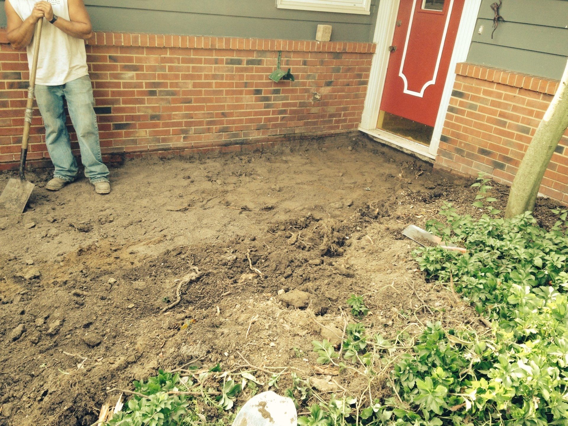 Freshly dug dirt beside a brick house with a red door; a person stands at the left edge.
