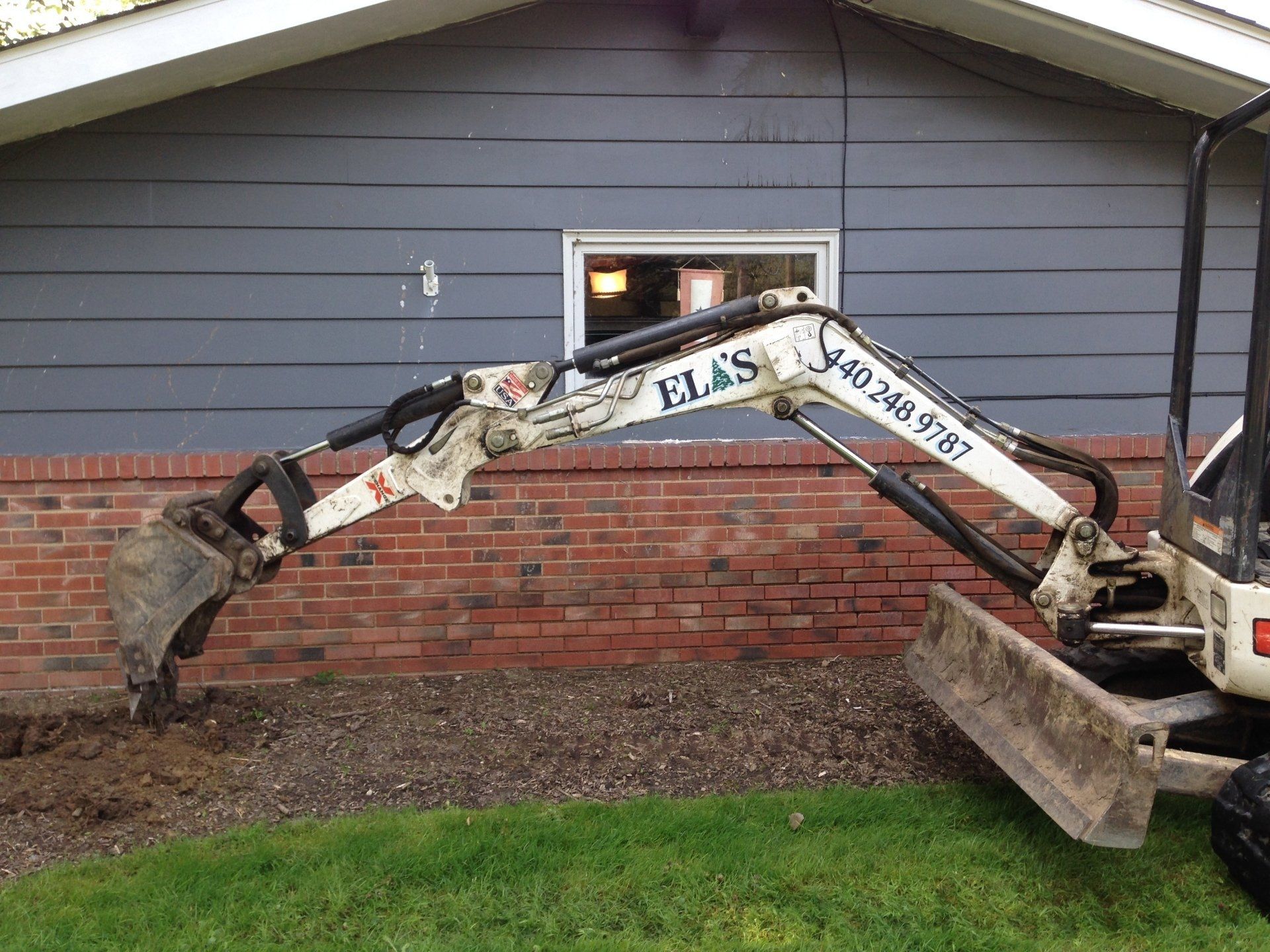 Excavator arm attached to a truck digging beside a brick house and window.