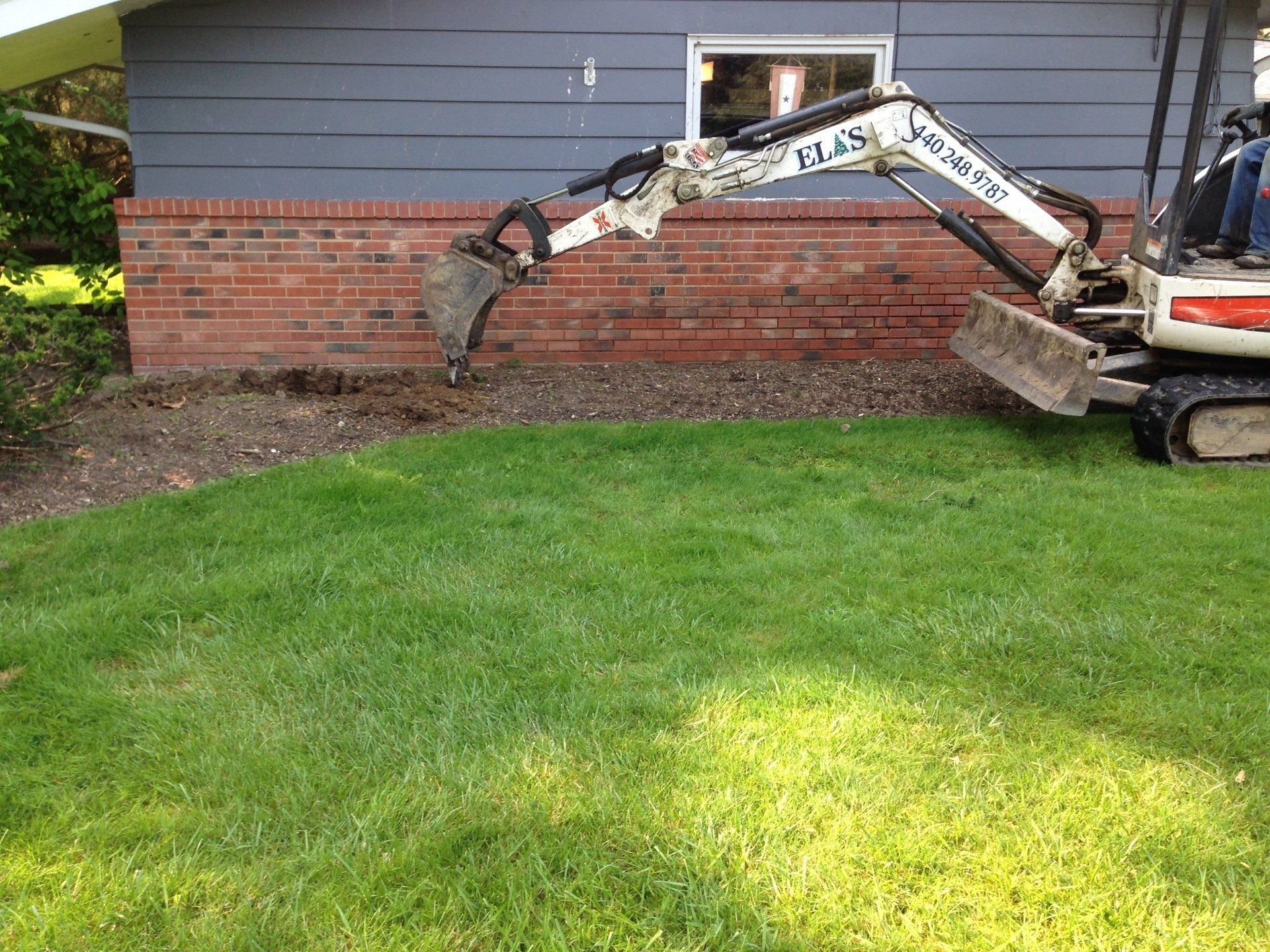 Mini excavator digging soil along a red brick foundation beside a gray house and green lawn