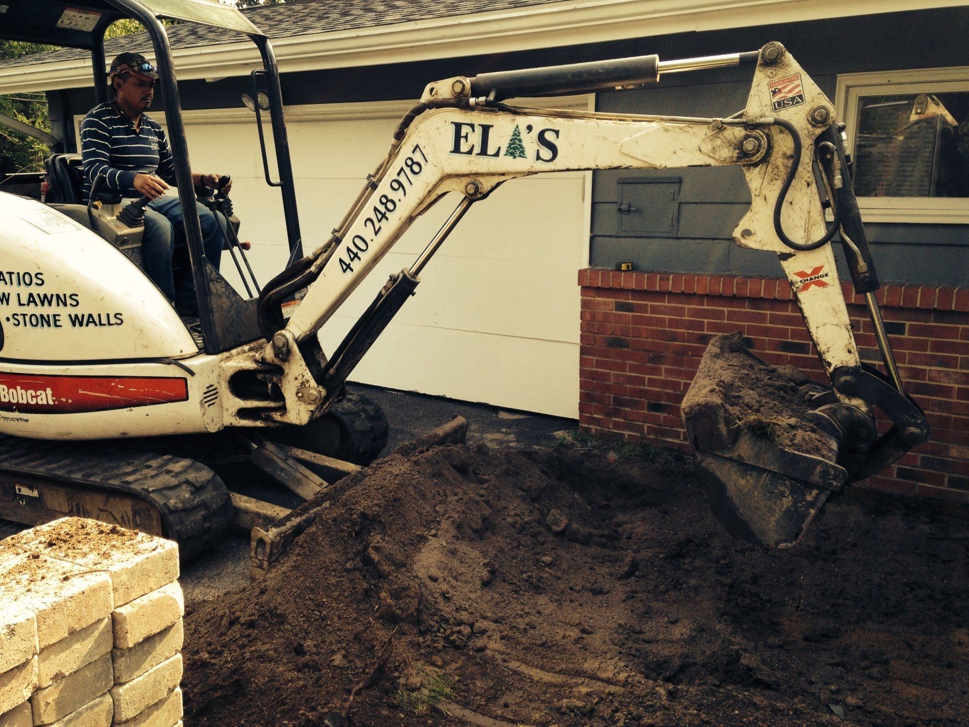 White excavator digging dirt beside a brick wall, with stacked bricks in the foreground.